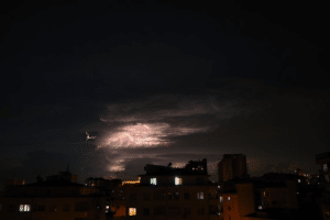 Nighttime storm clouds with lightning illuminating the sky over residential buildings, indicating severe weather conditions.