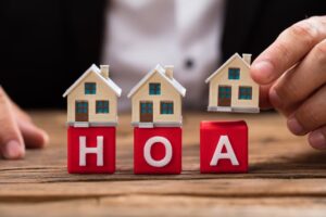 A businessperson's hand places a house model over red HOA blocks on a wooden desk.