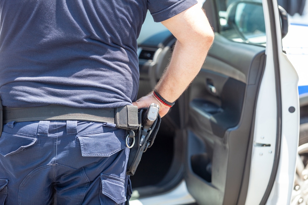 Police officer standing by a patrol car with hand on holstered firearm.